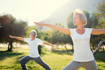 Mature couple shown in warrior 1 yoga pose in outside green space.